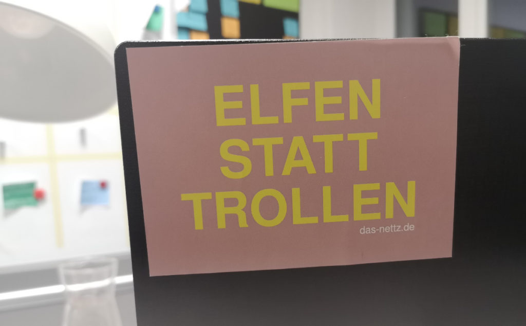 Man siehr den Aufkleber von "Das NETTZ" auf einem Bildschirm kleben. In gelber Schrift auf rosa steht dort "Elfen statt trollen".