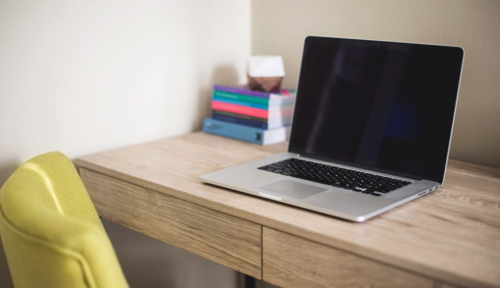 Ein Schreibtisch, auf dem ein Laptop und Bücher stehen. Vor dem Schreibtisch steht ein gelber Stuhl.