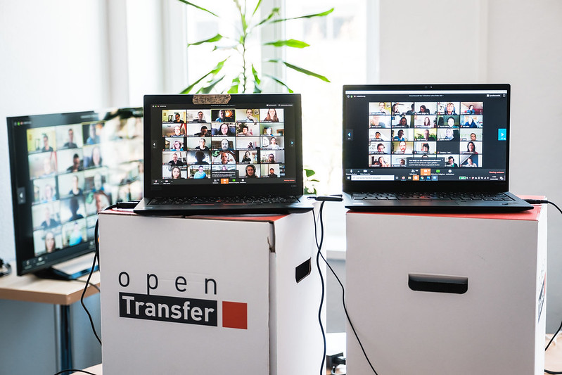 Zwei Laptops mit Kacheln eines vollen Zoom-Raums stehen auf Hockern von openTransfer. Im Hintergrund ein weiterer Monitor mit einem weiteren Zoom-Raum und eine Grünpflanze.