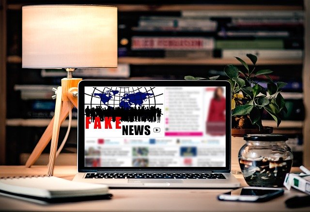 Auf einem Schreibtisch steht ein Lapttop, auf dem eine Nachrichtenseite zu sehen ist, auf der "Fake News" steht. drumhrum: Handy, Lampe, Bücherregal.