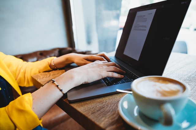 Man sieht Hände auf einer Tastatur eines Laptops, in dem ein Textdokument geöffnet ist. daneben auf dem Tisch steht eine Tasse Kaffee oder Cappuccino.
