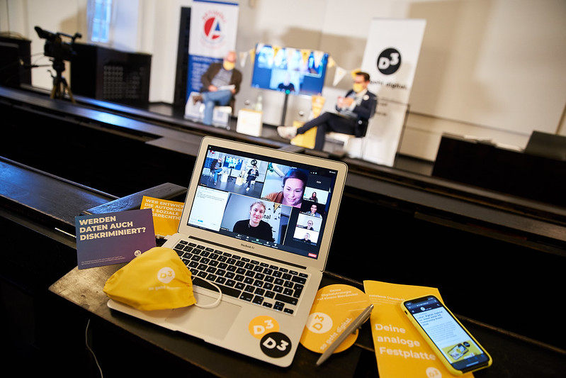 Im Vordergrund sieht man das D3-Team in einem Zoom-Call auf kleinen Kacheln auf einem Bildschirm. Der Laptop steht am oberen Rand eines Hörsaals, um ihn drapiert D3-Masken, Notizbücher und Sticker in gelb. Unten im Hörsaal sieht man verschwommen die Moderatoren Tom Leppert und Tomislav Perisic vor den Rollups von Aktivoli und D3 - so geht digital.